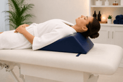 A woman in a white robe lies on a white massage table, using a dark blue wedge pillow for support in a spa-like room.