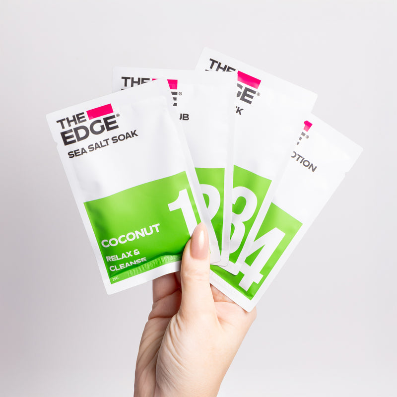 Hand holding three packets of 'The Edge' sea salt soak with coconut flavor on a light gray background.